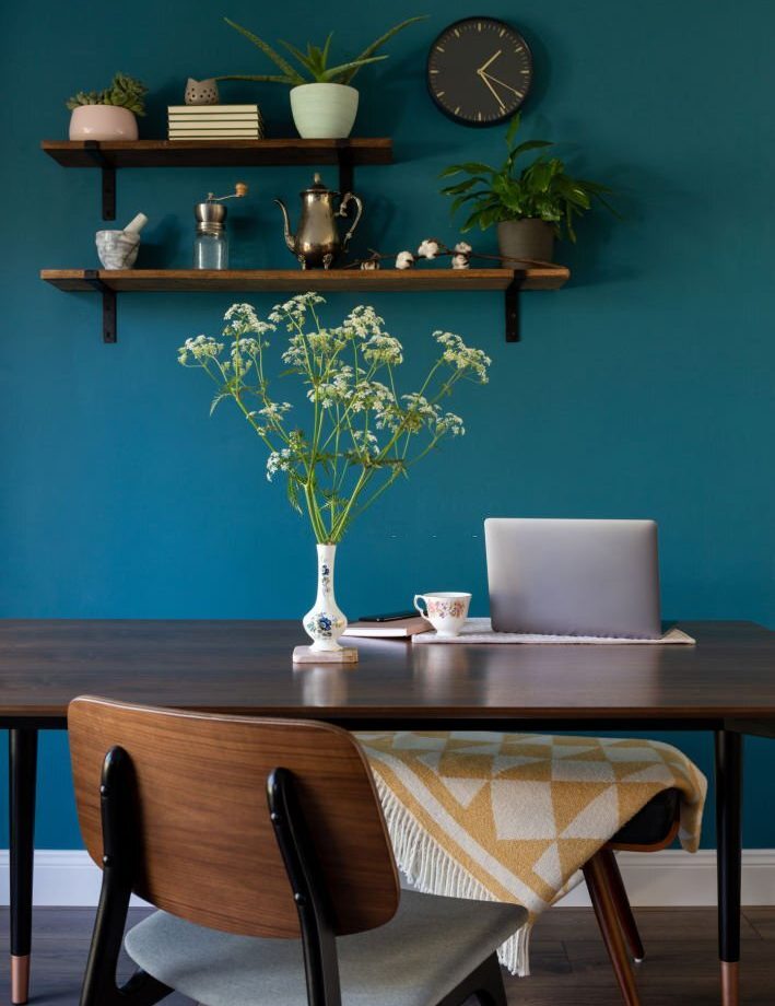 An open laptop at a kitchen table with a vintage vase of cow parsley wild flowers. Snug blanket with geometric print on a bench seat at dark wooden table. Modern deep petrol blue colour paint on the wall. The new home office, mobile professionals not just for freelancers.